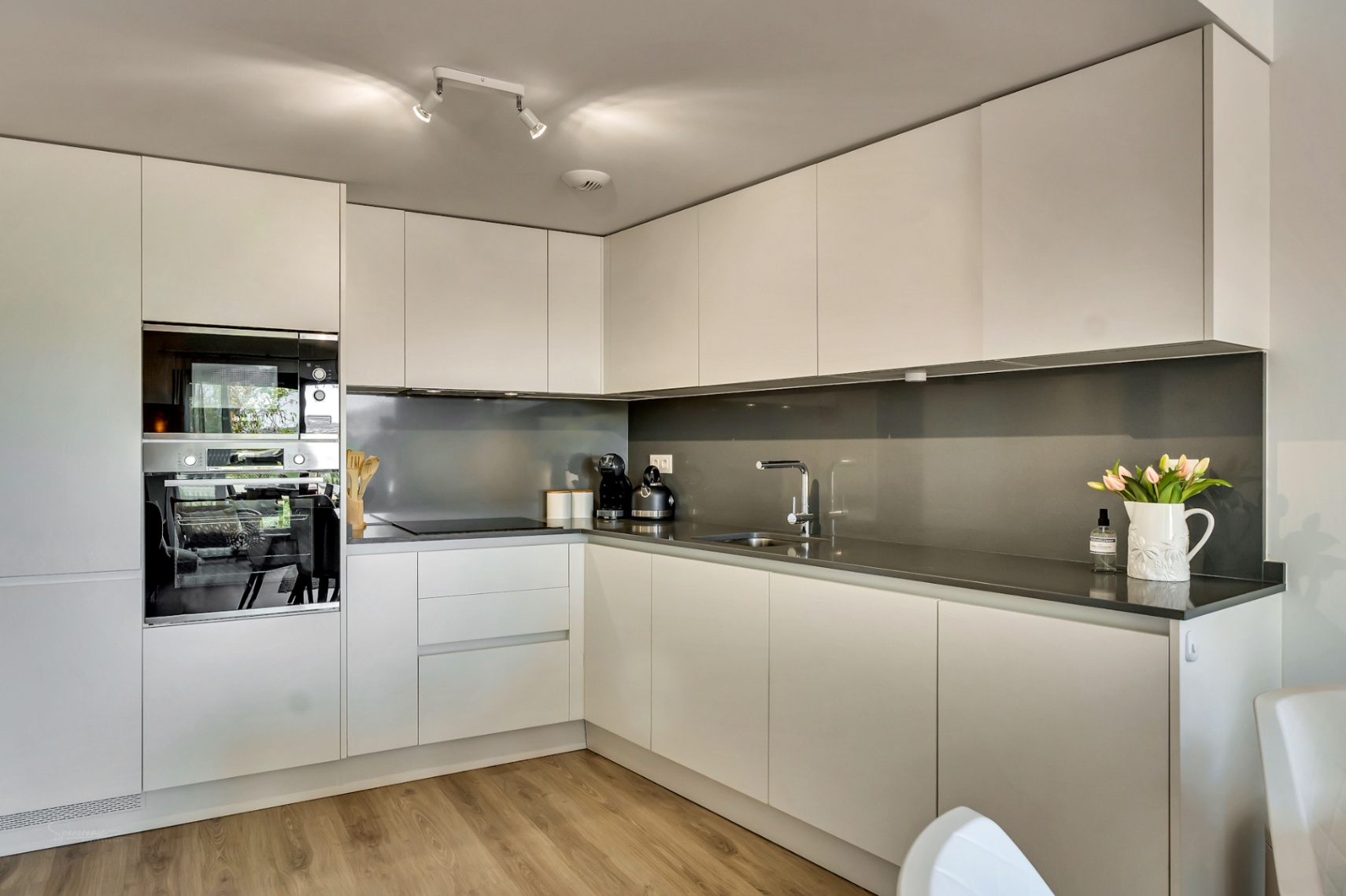 Modern L-shaped kitchen featuring handleless white cabinetry, dark grey stone countertops, and integrated appliances. Wood-effect flooring complements the sleek aesthetic.