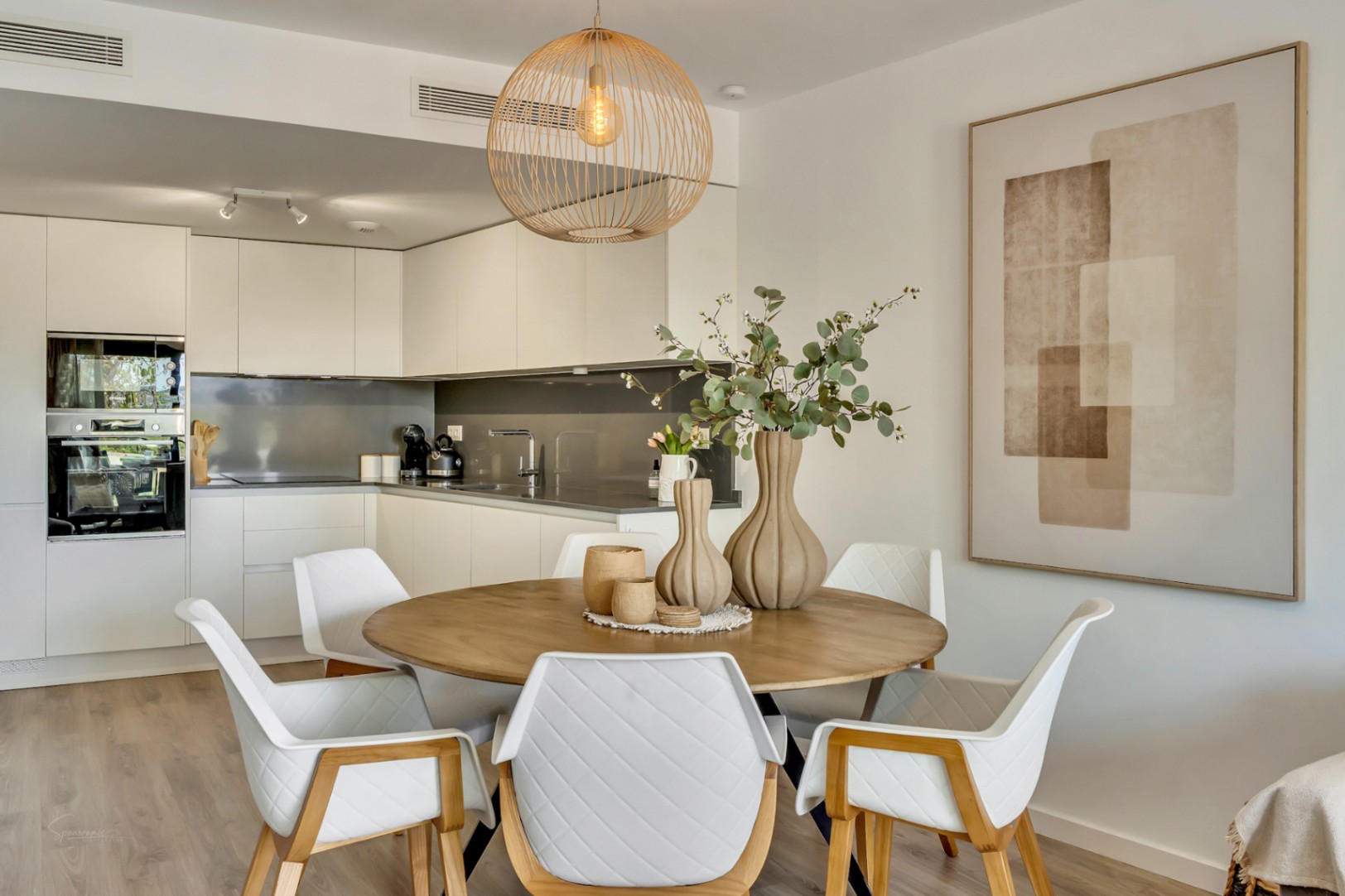 Open-concept dining area featuring a round wooden table, quilted chairs, and a rattan pendant light, seamlessly connected to a modern white kitchen.