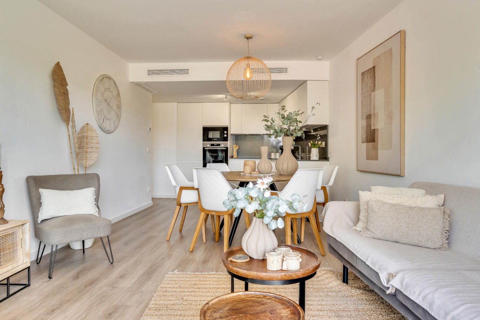 Open-concept living and dining area featuring light wood flooring, contemporary neutral furniture, and a modern white kitchen with integrated appliances and a dark backsplash.