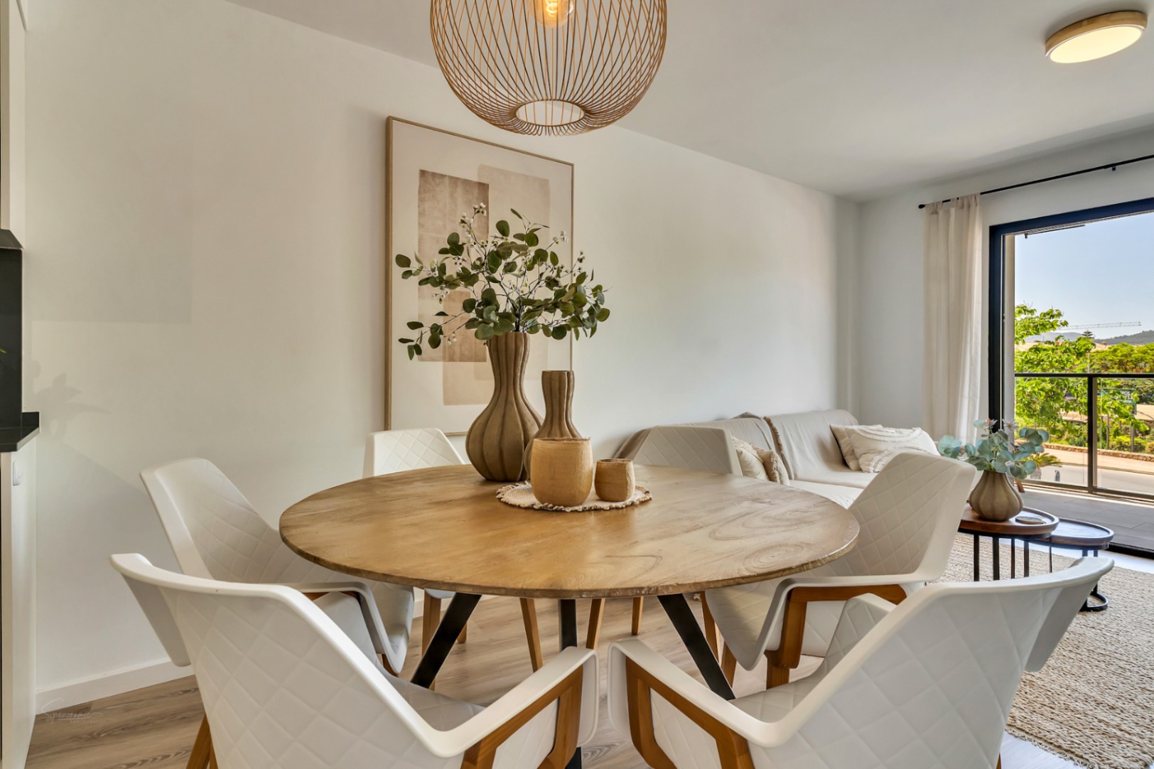 Open-plan dining area featuring a round wooden table, designer quilted chairs, and a rattan pendant light, connected to a bright living space with balcony access.