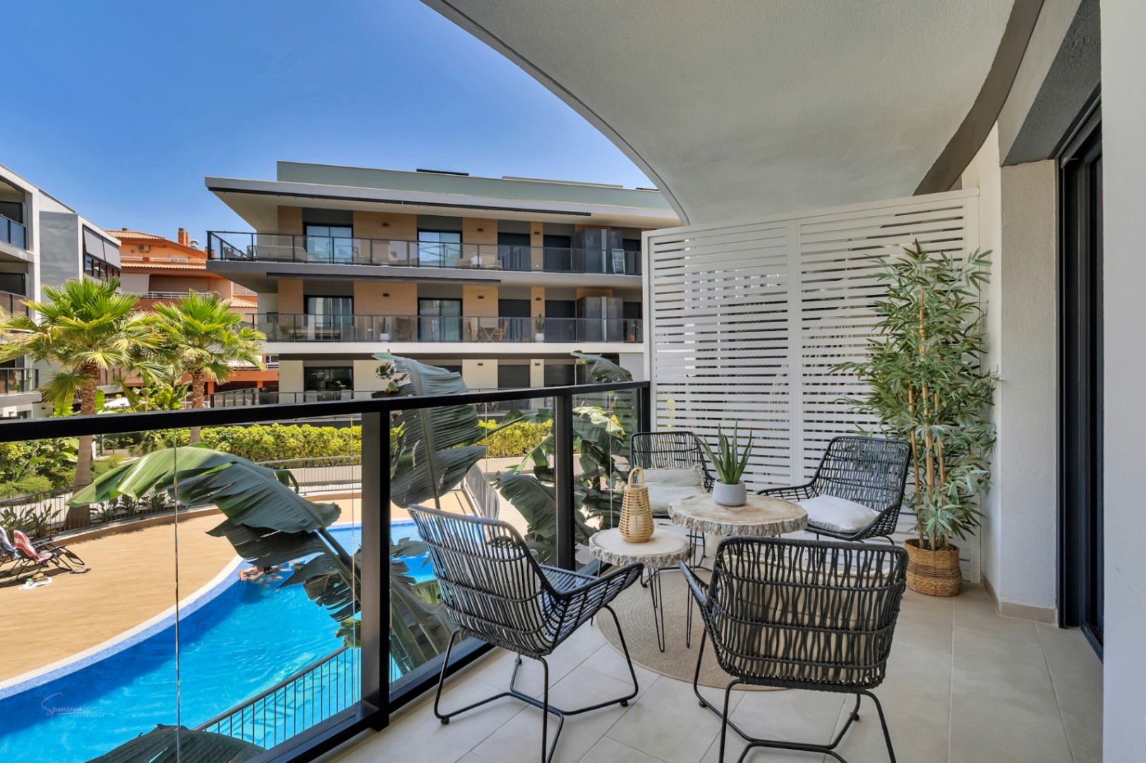 Private terrace featuring a glass balustrade, contemporary outdoor seating, and direct views over the communal pool and landscaped Mediterranean gardens.