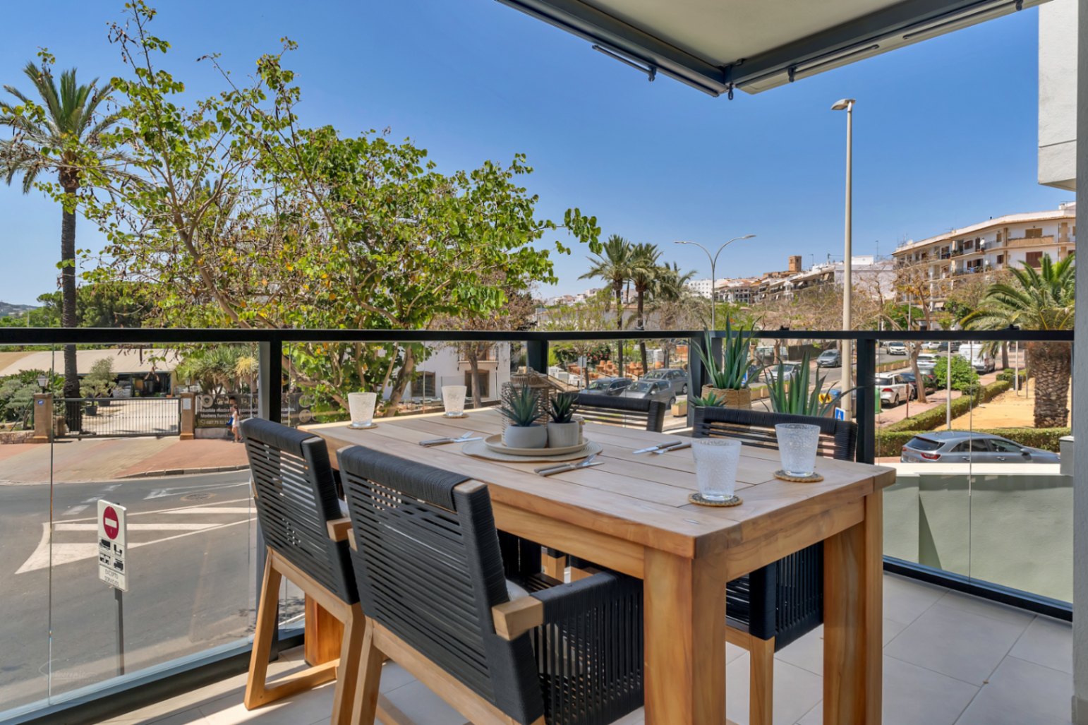 Private terrace featuring a wooden dining set with woven chairs, glass balustrades, and open views of the surrounding palm trees and urban landscape.