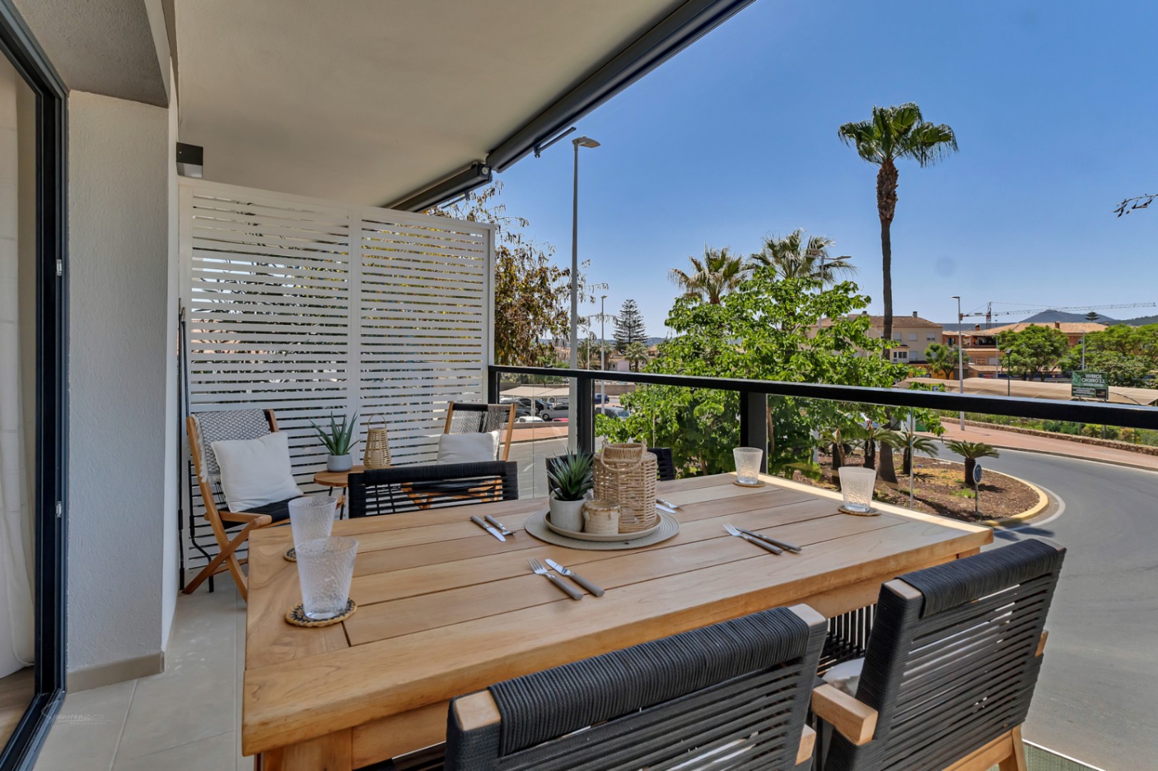 Private terrace featuring a wooden dining table, woven cord chairs, and glass railings overlooking palm trees and the surrounding urban landscape.