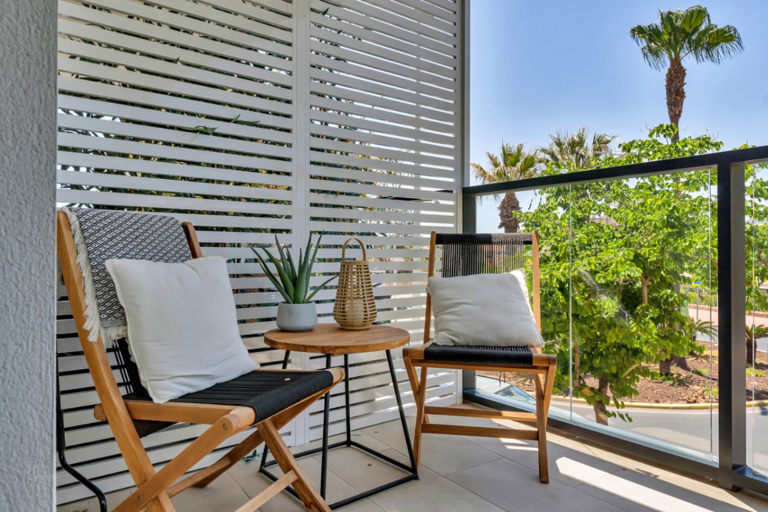 Private terrace featuring wooden lounge chairs, a slatted privacy screen, and a glass railing overlooking Mediterranean greenery and palm trees.