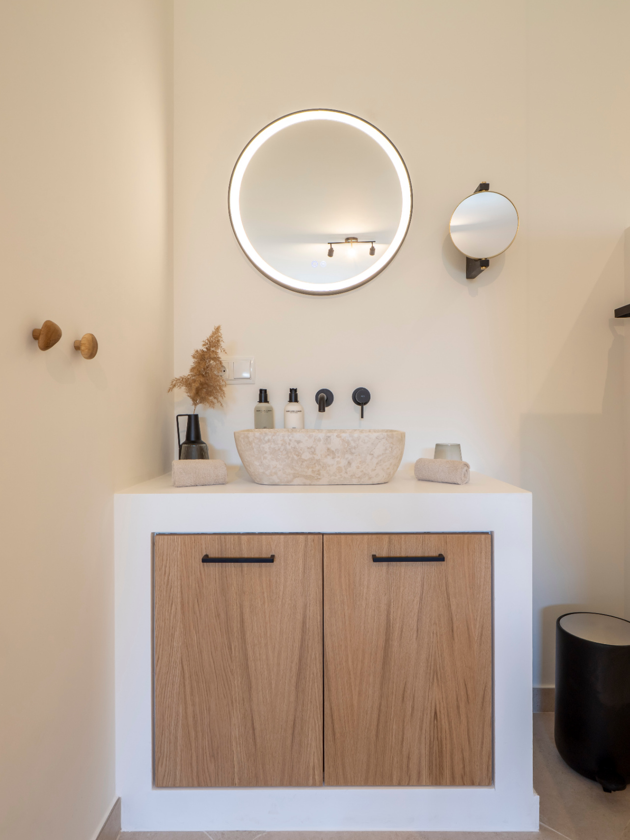 Bathroom detail with natural stone sink, backlit circular mirror, and light wood cabinet with black handles.