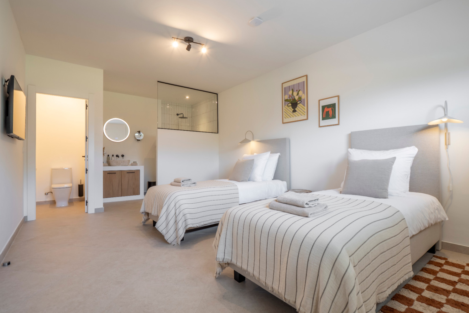 Bright bedroom featuring twin beds, en-suite bathroom with glass-enclosed shower, and light wood vanity.