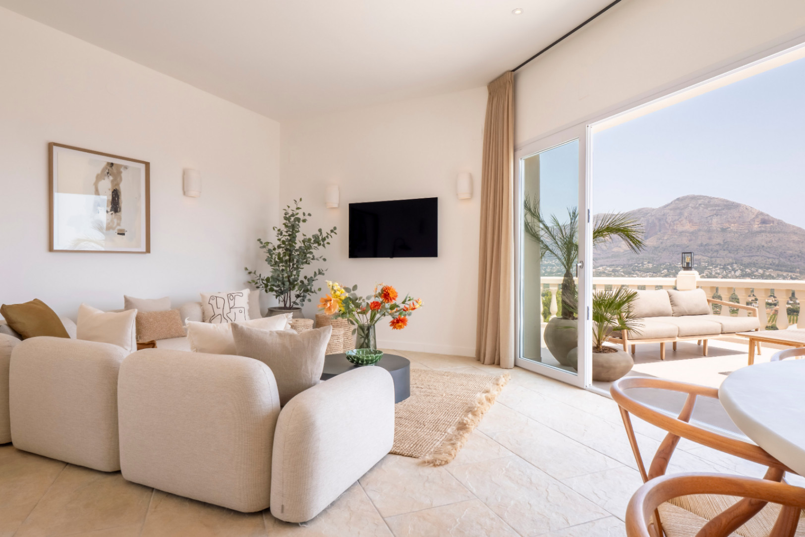 Bright living room with terrace access. Beige modular sofa, TV, and floral decor. Mountain views in Jávea.