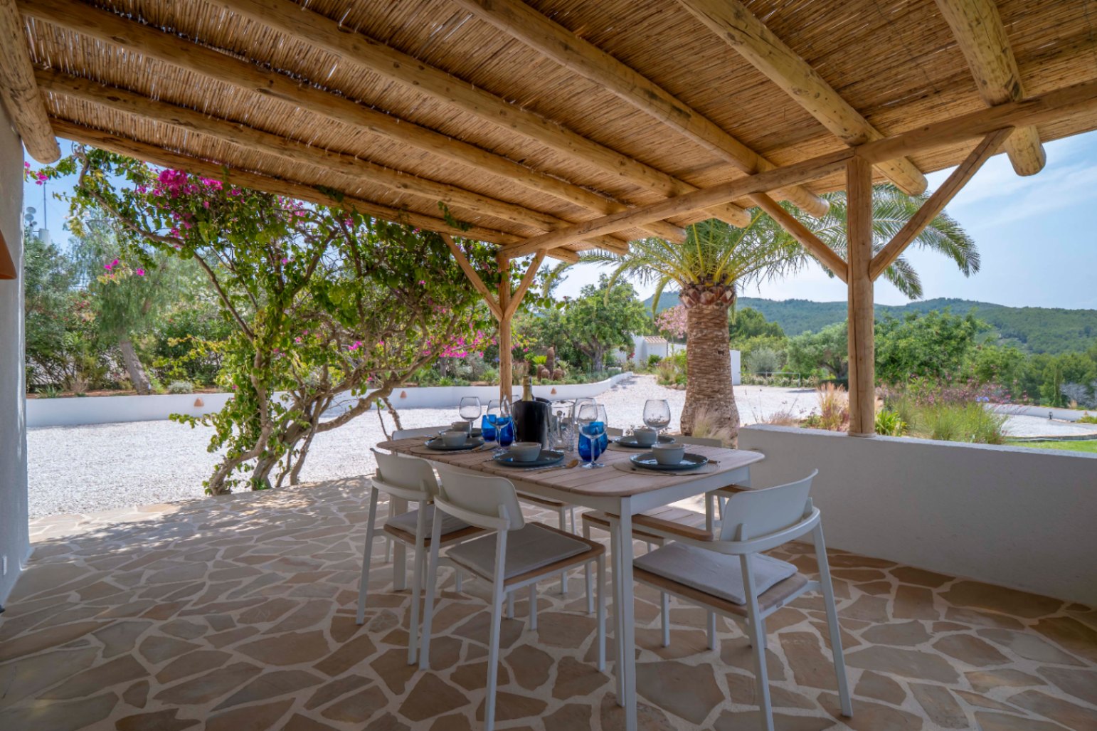 Covered outdoor dining area with thatched roof, irregular stone floor, and views of the Mediterranean garden.