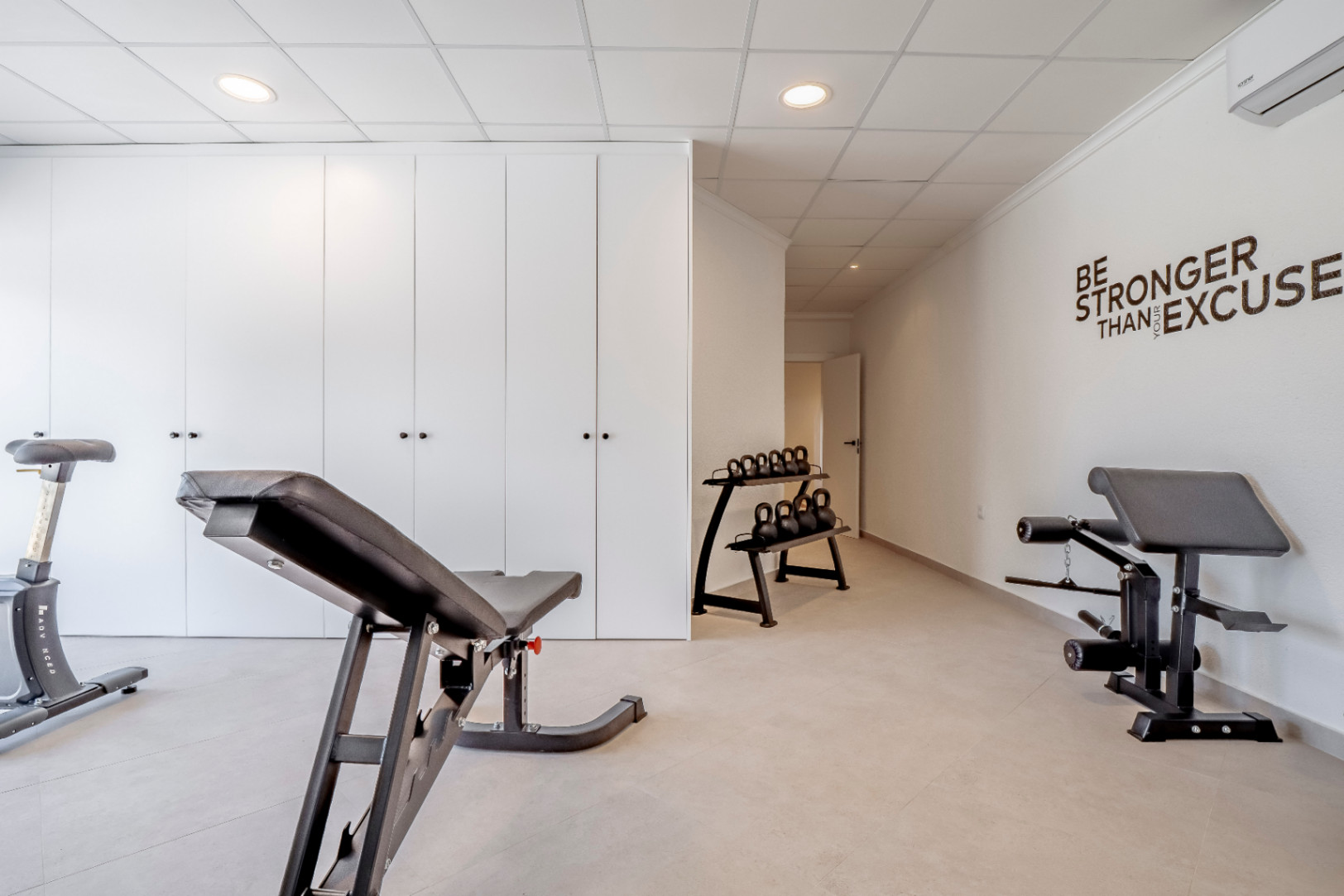 Gym with ceramic floors, white walls and built-in wardrobes. Training equipment with weights and bench.