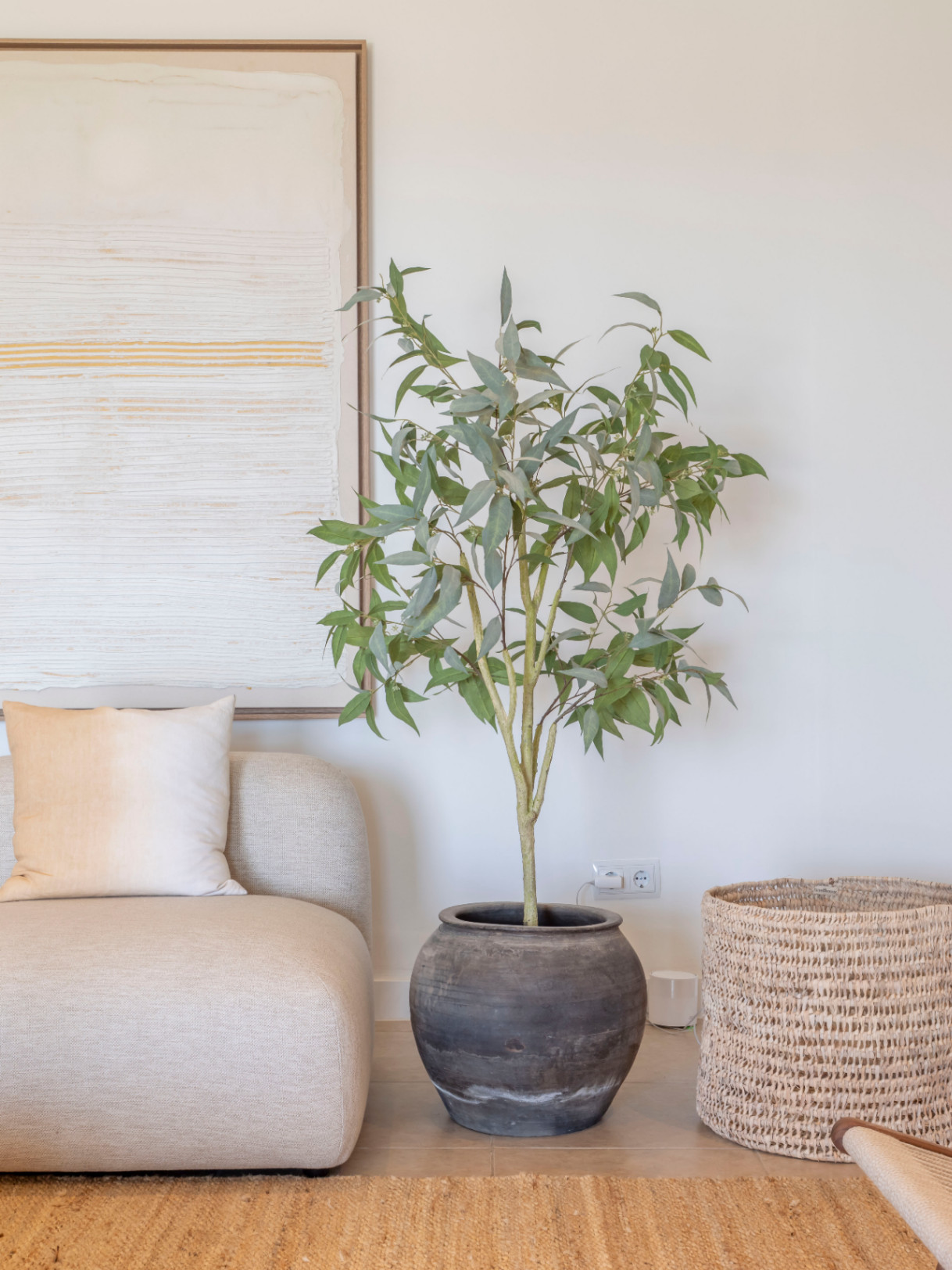 Living room detail with beige sofa, potted ceramic plant, and wicker basket on a jute rug.