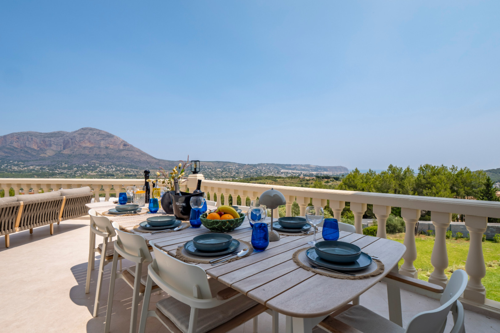 Outdoor dining area featuring a wooden table and panoramic mountain & coastal views from the terrace.