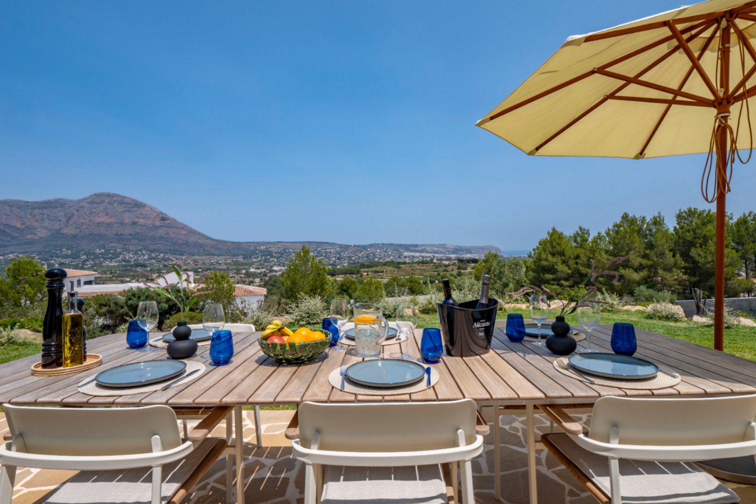 Outdoor dining area with wooden table, blue tableware, and panoramic mountain views in Jávea.
