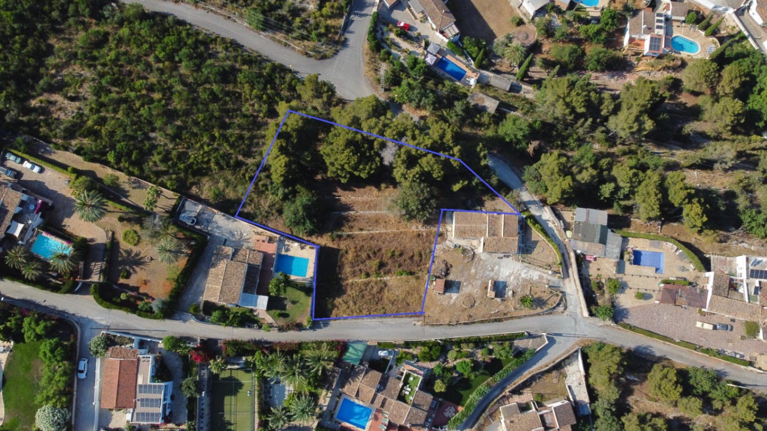 Aerial drone view showing the blue-outlined 1,908 sqm plot in Jávea, featuring terraced land, existing structures, and mature trees within a developed neighborhood.