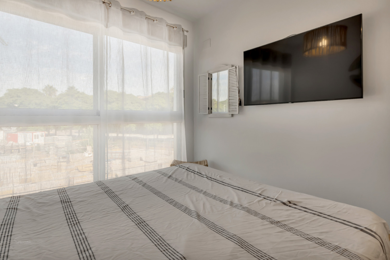 Bright bedroom featuring a large window with sheer curtains, a double bed with striped linens, and a wall-mounted flat-screen television.
