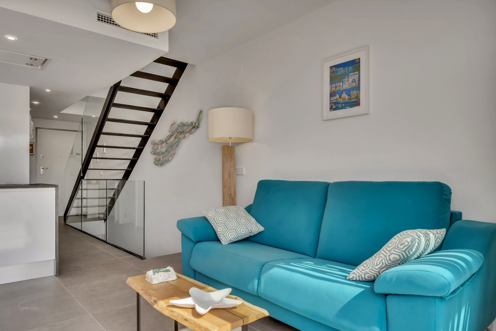 Bright living room featuring a turquoise sofa, natural wood coffee table, and a minimalist black metal staircase with glass railing. Grey porcelain tile flooring and open design.