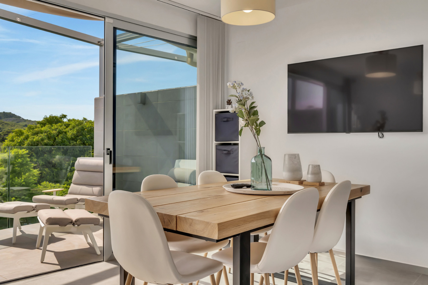Dining area featuring a solid wood table and white chairs, seamlessly connected to a terrace with lounge seating and views of the surrounding greenery.