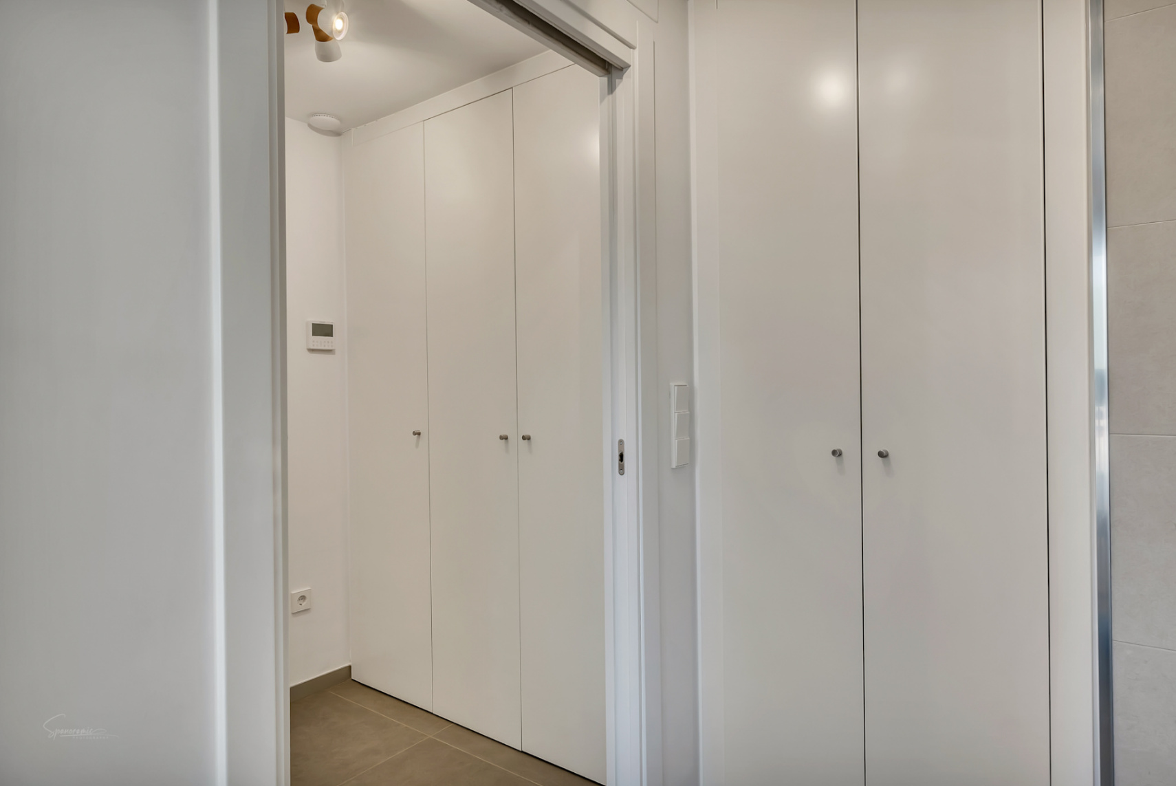 Floor-to-ceiling built-in wardrobes with a minimalist white finish and subtle metal handles, featuring integrated storage and a digital climate or alarm control panel.