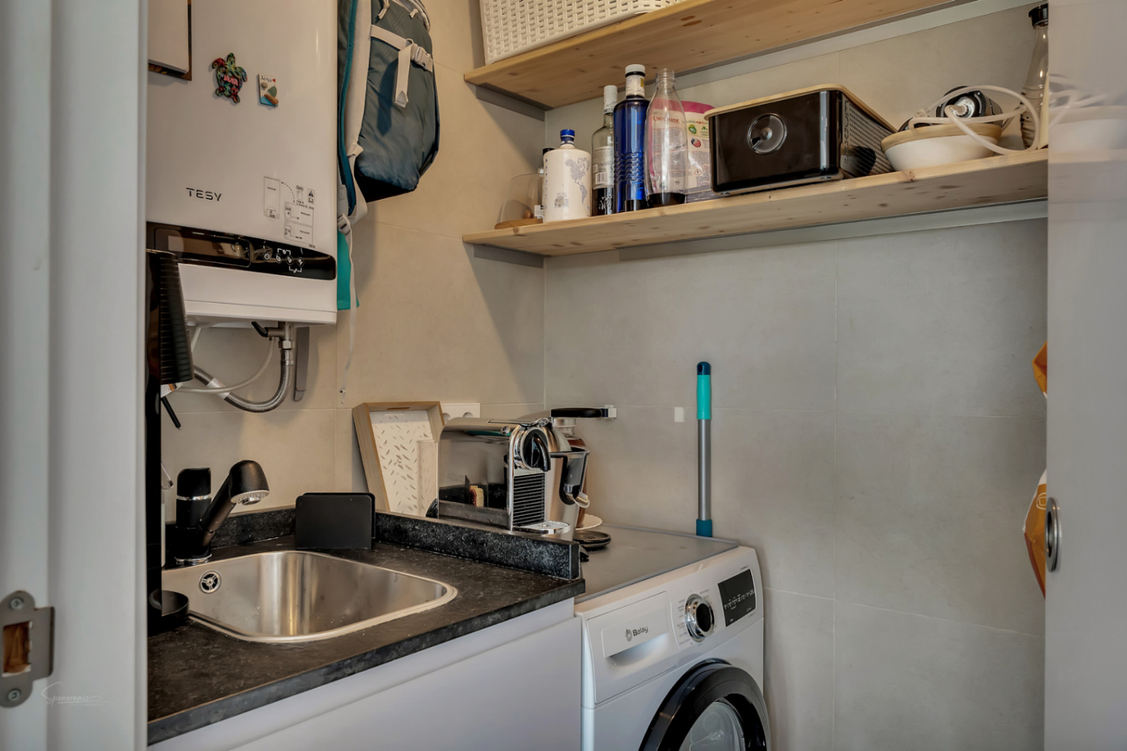 Functional laundry area equipped with a Balay washing machine, stainless steel sink, Tesy electric water heater, and wooden shelving.
