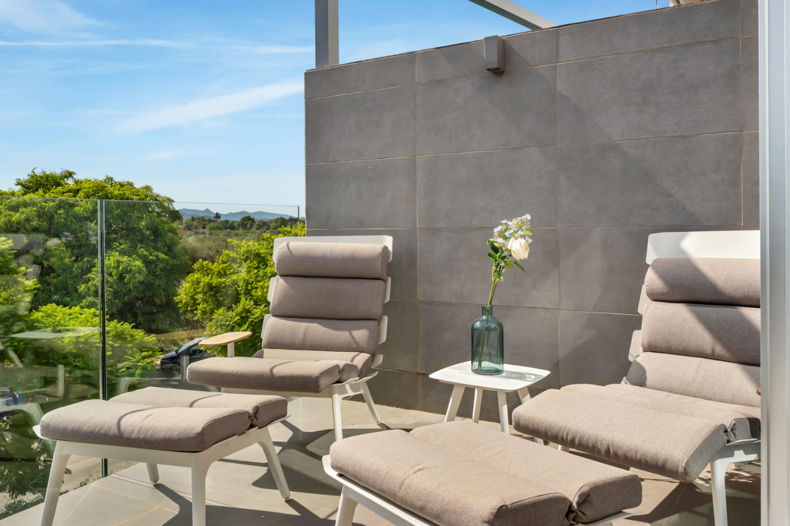Modern balcony featuring glass railings, grey porcelain tiling, and contemporary lounge seating. It offers unobstructed views of protected fields and distant mountains.