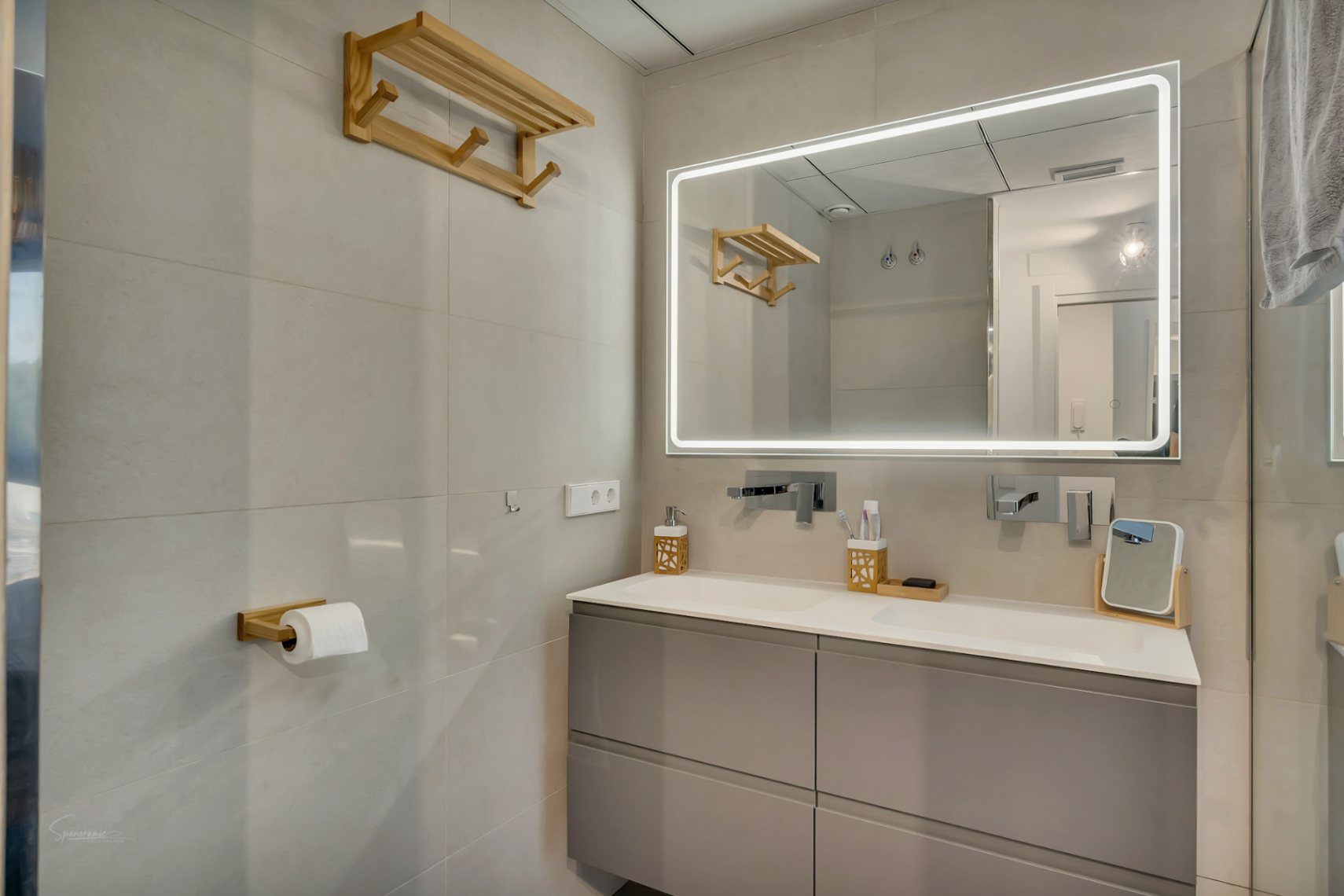 Modern bathroom featuring a grey floating double vanity, integrated twin sinks, and a large LED-backlit mirror. Wall-mounted chrome faucets and natural wood accents complete the space.