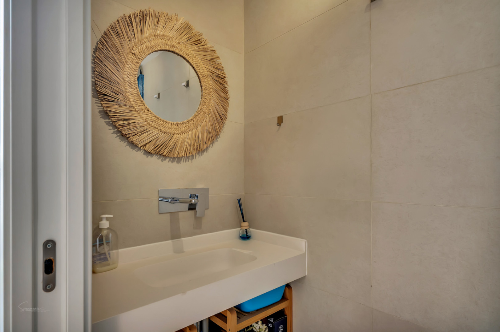 Modern bathroom featuring a white integrated sink, wall-mounted chrome faucet, and a decorative fiber-framed mirror against large-format sand-toned wall tiles.