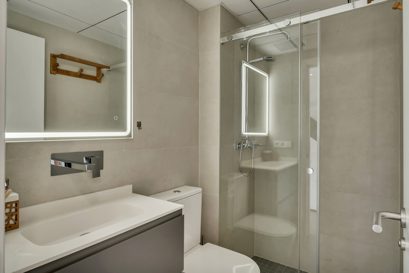 Modern bathroom featuring neutral tiles, a grey vanity, and a mirror with perimeter LED lighting next to a walk-in shower with sliding glass doors.