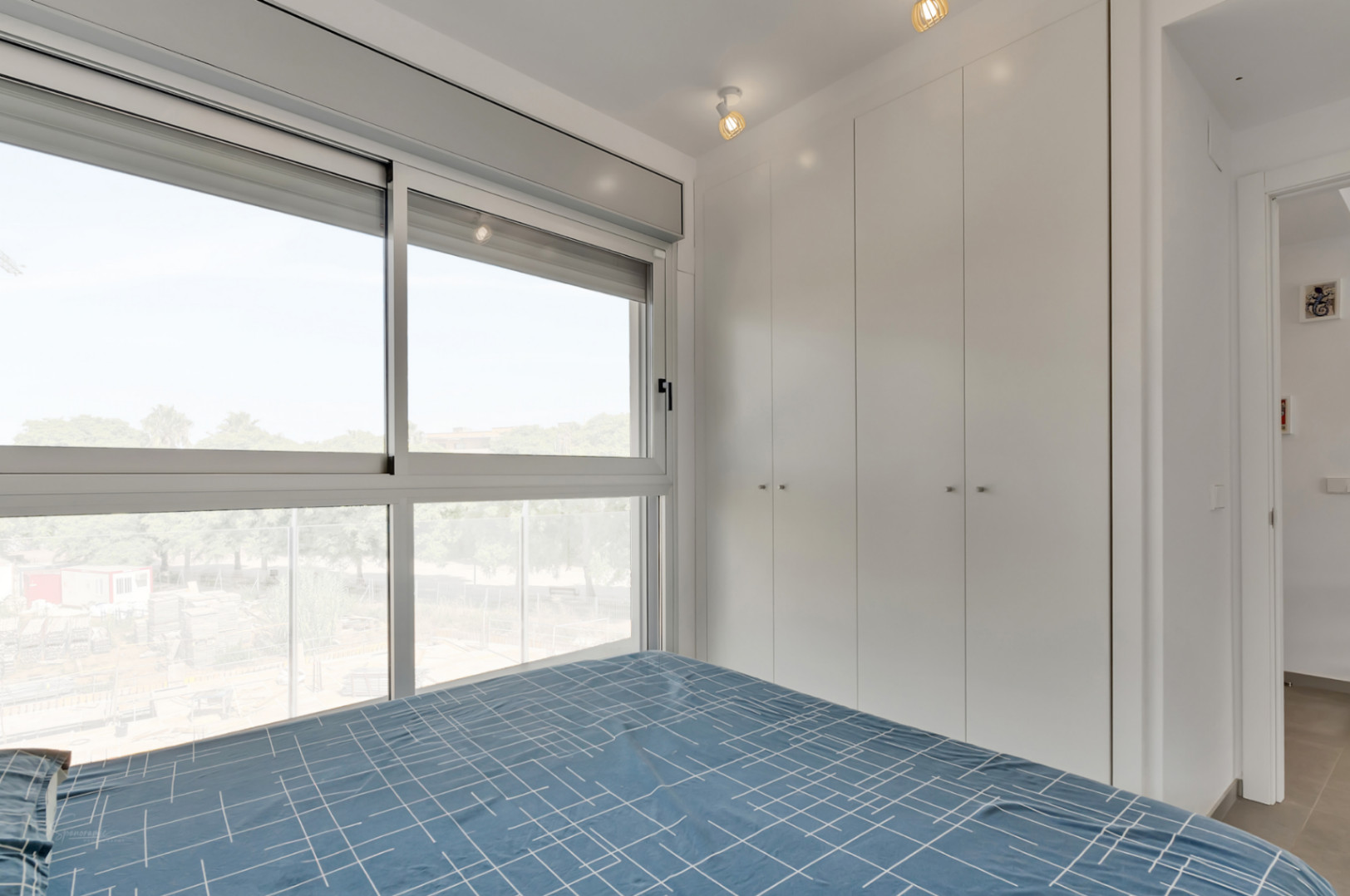 Modern bedroom featuring floor-to-ceiling white built-in wardrobes and a large window with motorized blinds providing ample natural light.