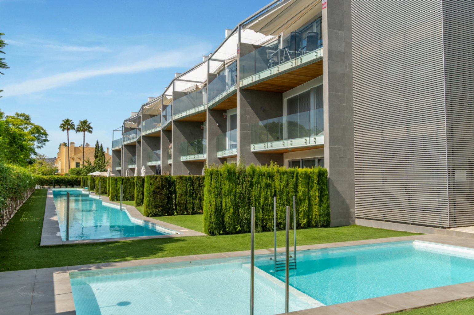 Modern building exterior featuring glass balconies with wood finishes, overlooking a long communal pool and manicured green areas.