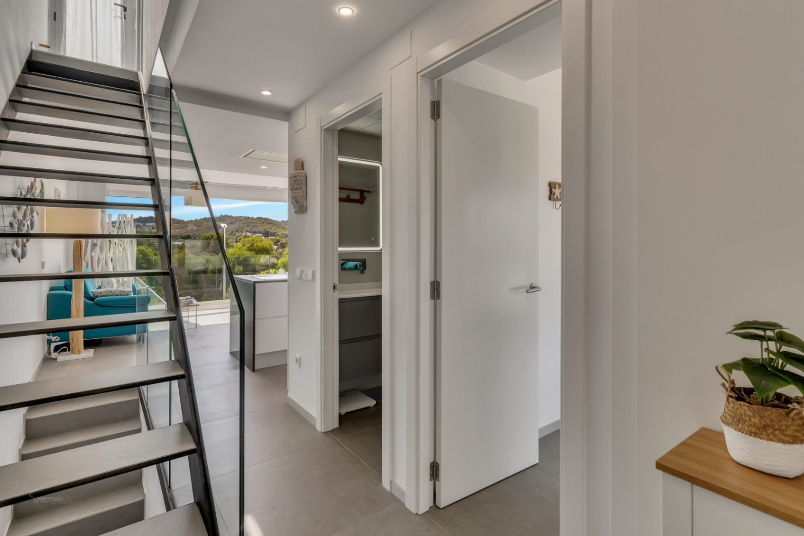 Modern hallway featuring an open-tread staircase with glass balustrade. Access to a contemporary bathroom with backlit mirror and views toward the bright living area.