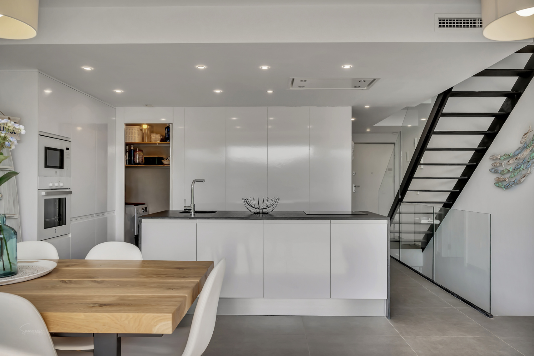 Modern minimalist kitchen featuring a central island with dark countertop, high-gloss cabinetry, and a black metal staircase with glass railings next to the wooden dining table.