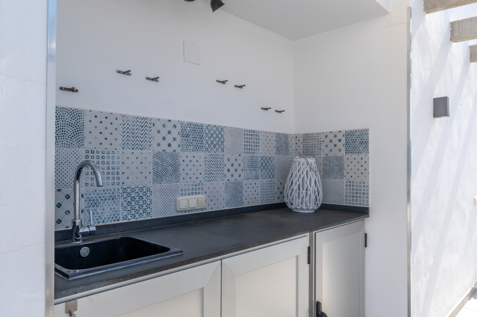 Outdoor summer kitchen featuring a dark countertop, integrated sink, and a decorative blue and white patterned tile backsplash.
