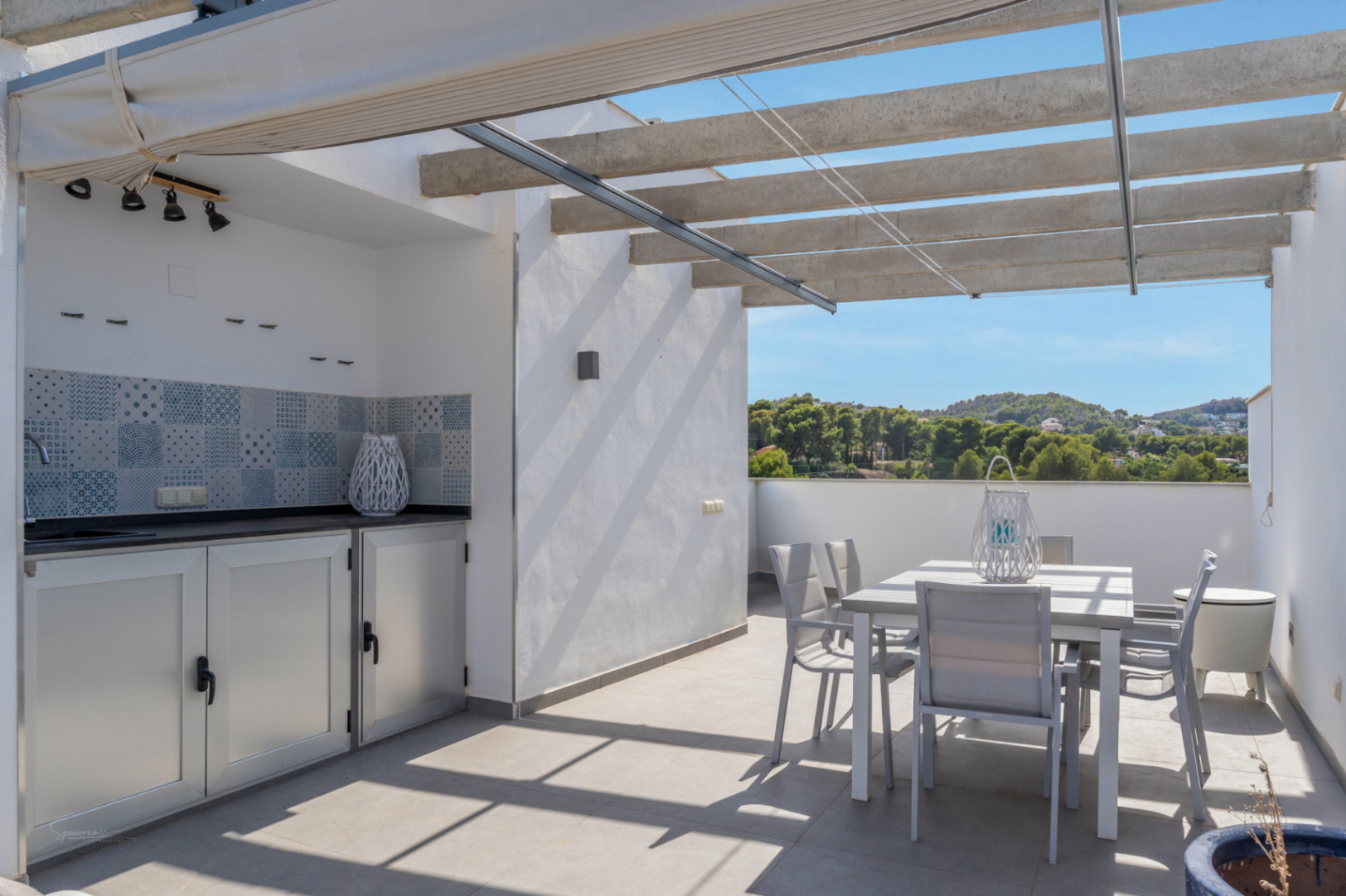 Private rooftop terrace featuring a summer kitchen with tiled backsplash, outdoor dining area, and concrete pergola with retractable awning overlooking green hills.