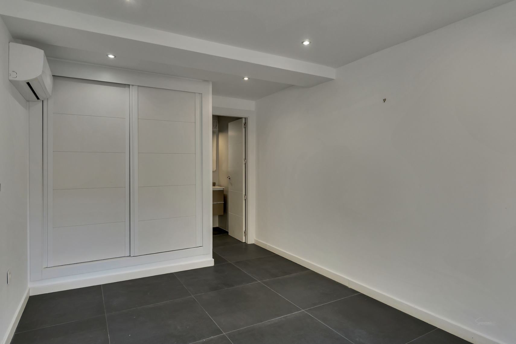 Bedroom featuring a large white built-in sliding wardrobe, dark tiled flooring, recessed lighting, and an integrated air conditioning unit next to the en-suite.