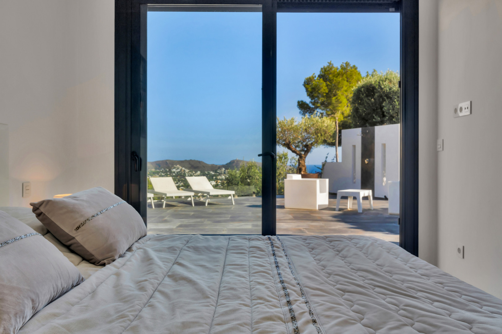 Bedroom featuring large sliding glass doors that open onto a stone-tiled terrace with sun loungers, Mediterranean trees, and distant sea views.