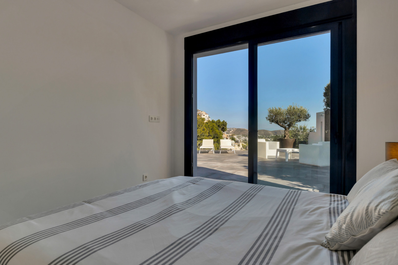 Bedroom with direct terrace access through large sliding doors. The view overlooks the outdoor lounge area, a potted olive tree, and the surrounding hills.