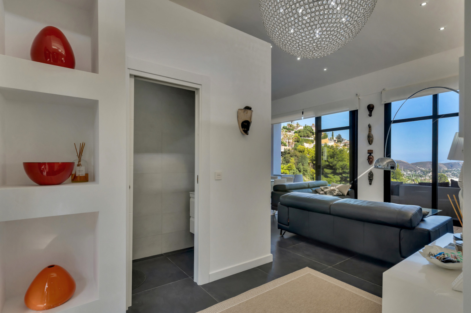 Bright entrance featuring recessed wall niches and dark tiling, leading to a living area with floor-to-ceiling windows and a designer crystal chandelier.