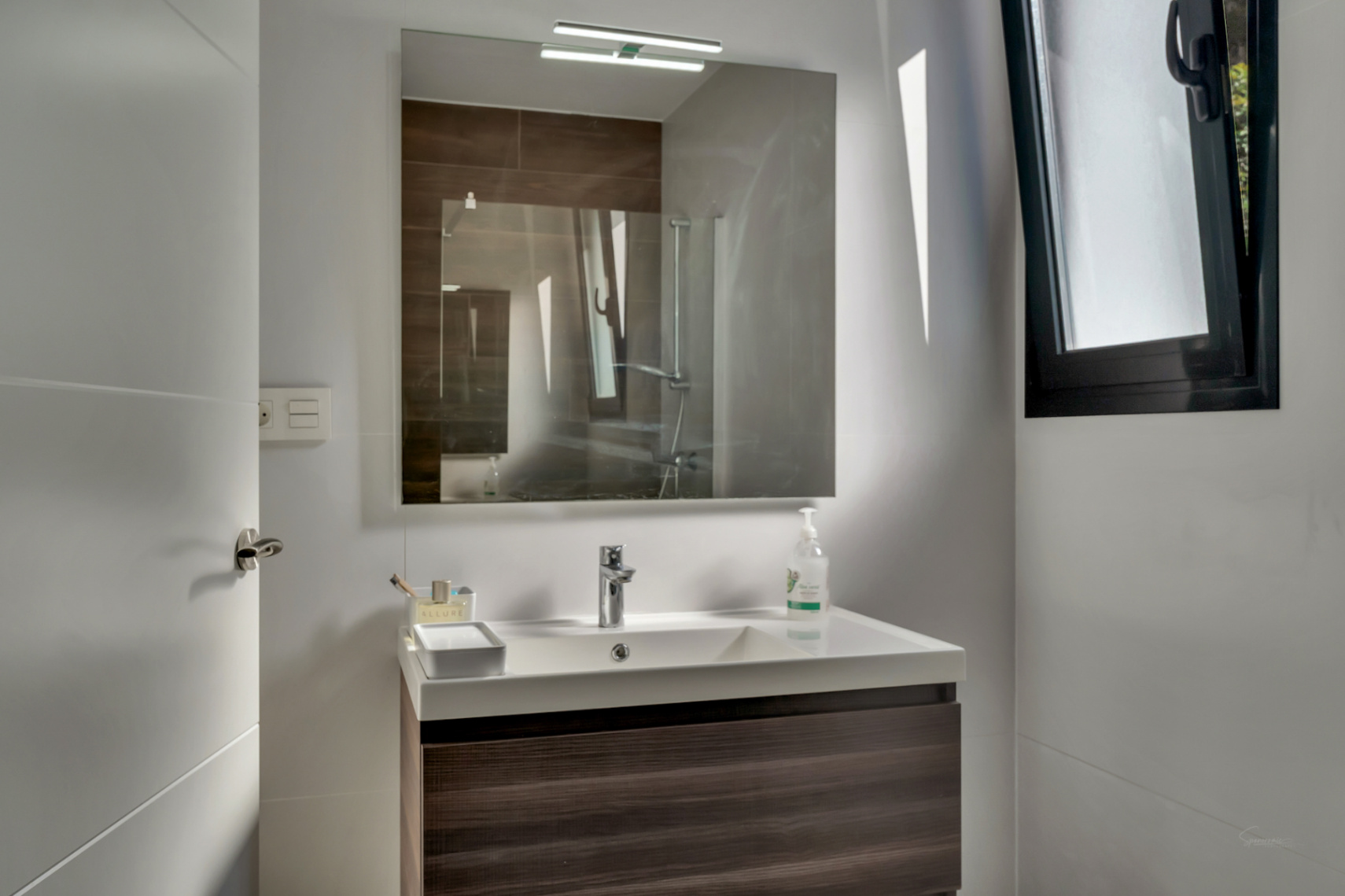 Contemporary bathroom with a dark wood floating vanity, white integrated sink, and a large mirror with top lighting. Features a black-framed window.