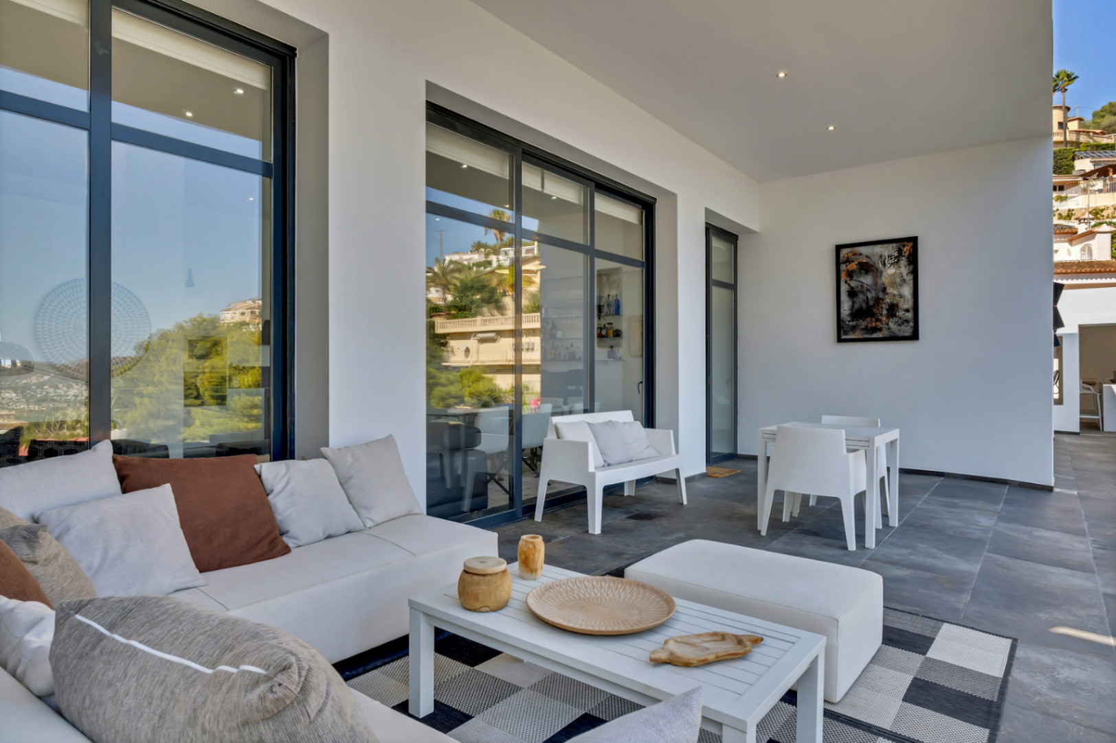 Covered terrace featuring a modern lounge area, grey tiled flooring, and large black-framed glass doors reflecting the coastal landscape.