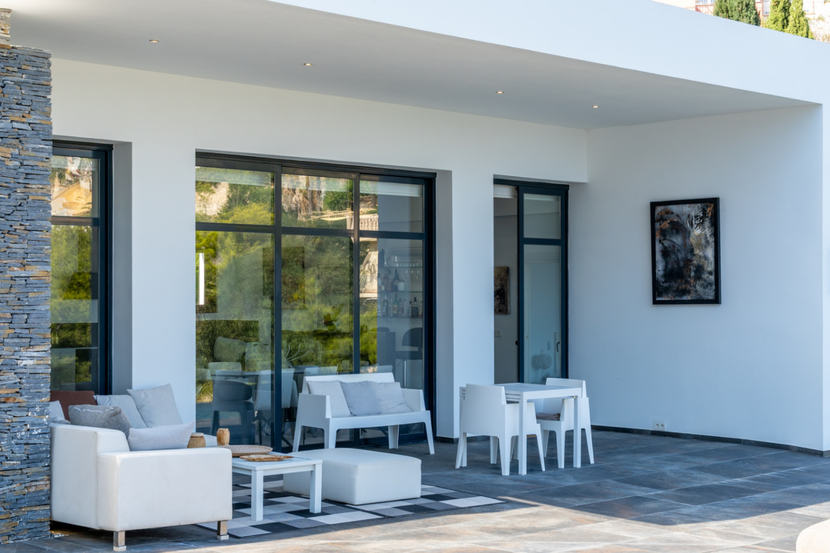 Covered terrace featuring dark porcelain flooring, natural stone cladding, and minimalist white outdoor furniture integrated with large sliding glass doors.