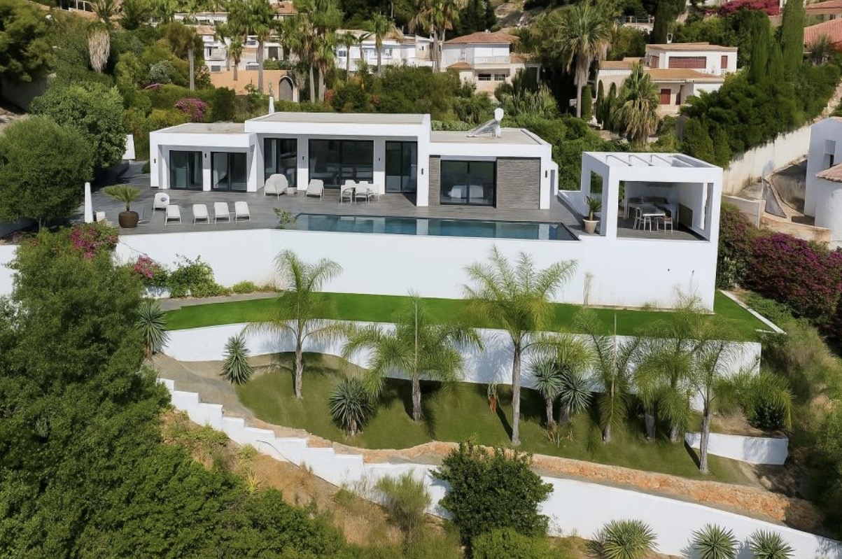 Elevated view of the modern white villa featuring a linear pool, expansive sun terrace, and tiered Mediterranean gardens with palm trees and manicured lawns.