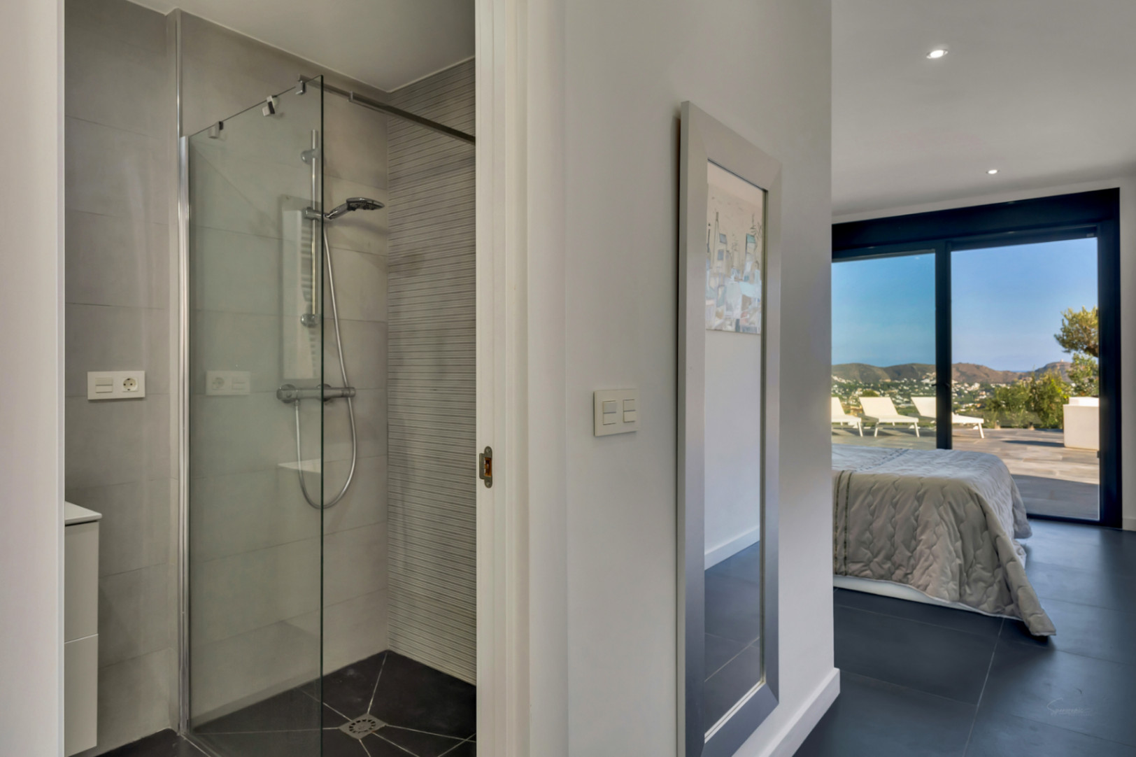 Ensuite bathroom featuring a walk-in glass shower and grey tiling, opening into a bedroom with large sliding doors and panoramic outdoor views.