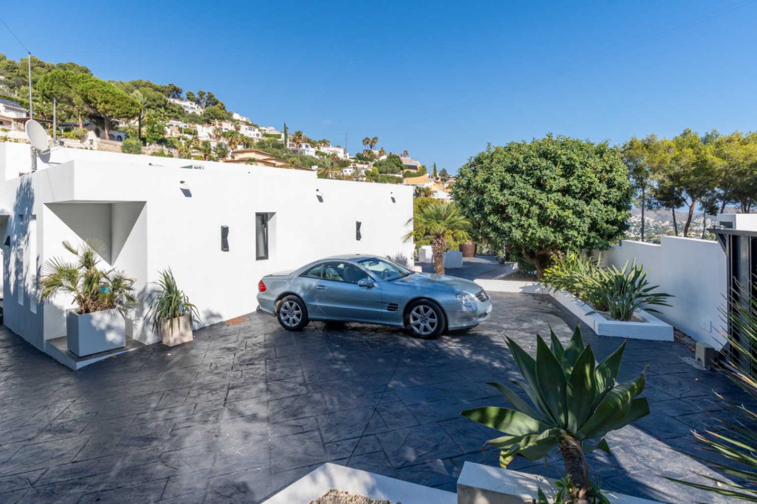 Entrance area and parking featuring dark stamped concrete flooring, a minimalist white facade, and landscaped planters with Mediterranean vegetation.