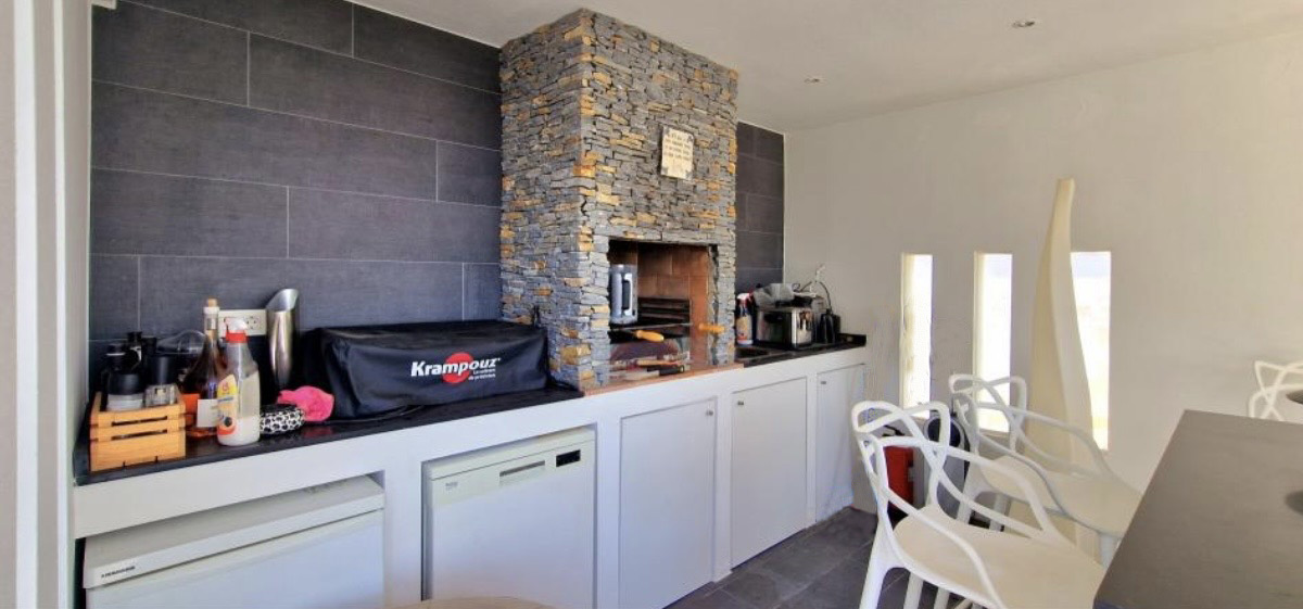 Fully equipped summer kitchen featuring a natural stone barbecue, dark grey tiled walls, and integrated appliances under a sleek black countertop.