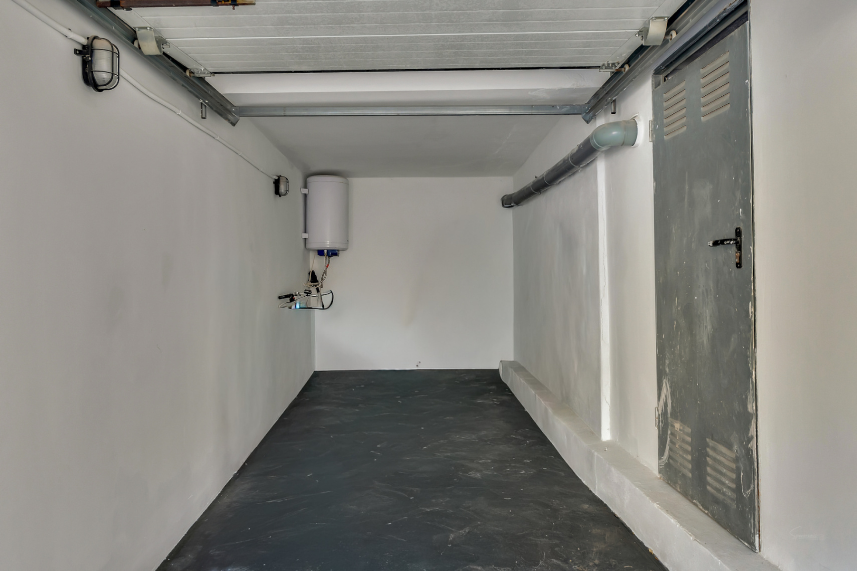 Functional garage and storage space featuring dark flooring, white walls, an electric water heater, and a lateral metal door.