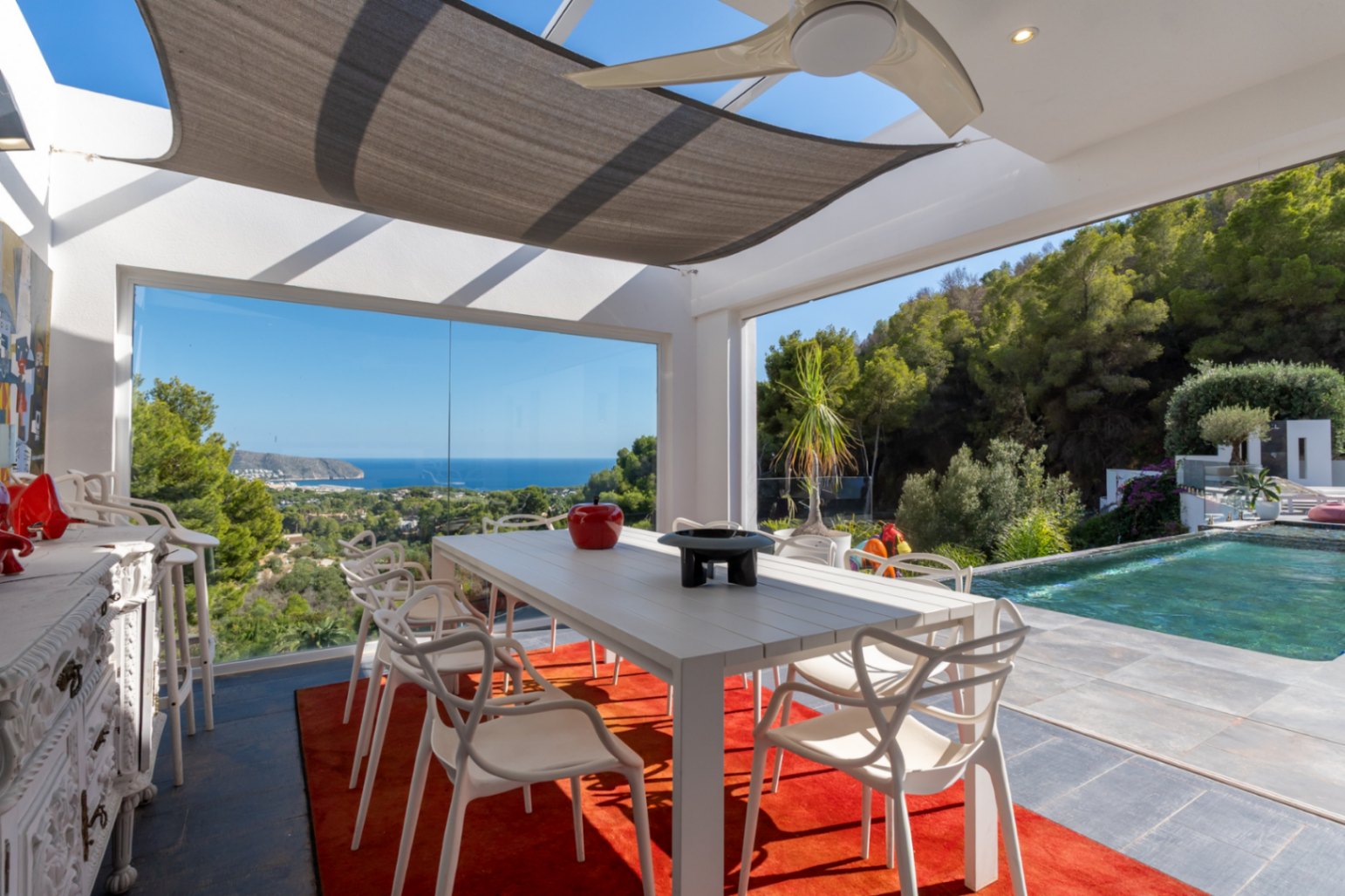 Glazed outdoor dining area featuring designer seating, a sunshade sail, and panoramic Mediterranean views adjacent to the infinity pool and lush hillside.