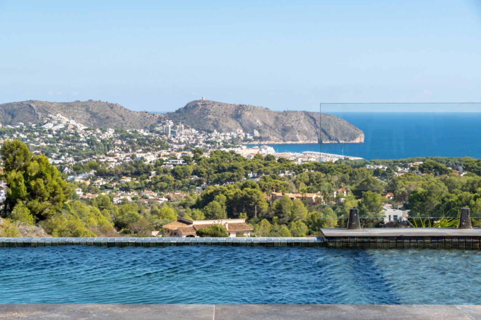 Infinity pool with blue mosaic tiling and a glass balustrade, featuring panoramic views of the Mediterranean Sea, Cap d'Or, and the lush coastal landscape.