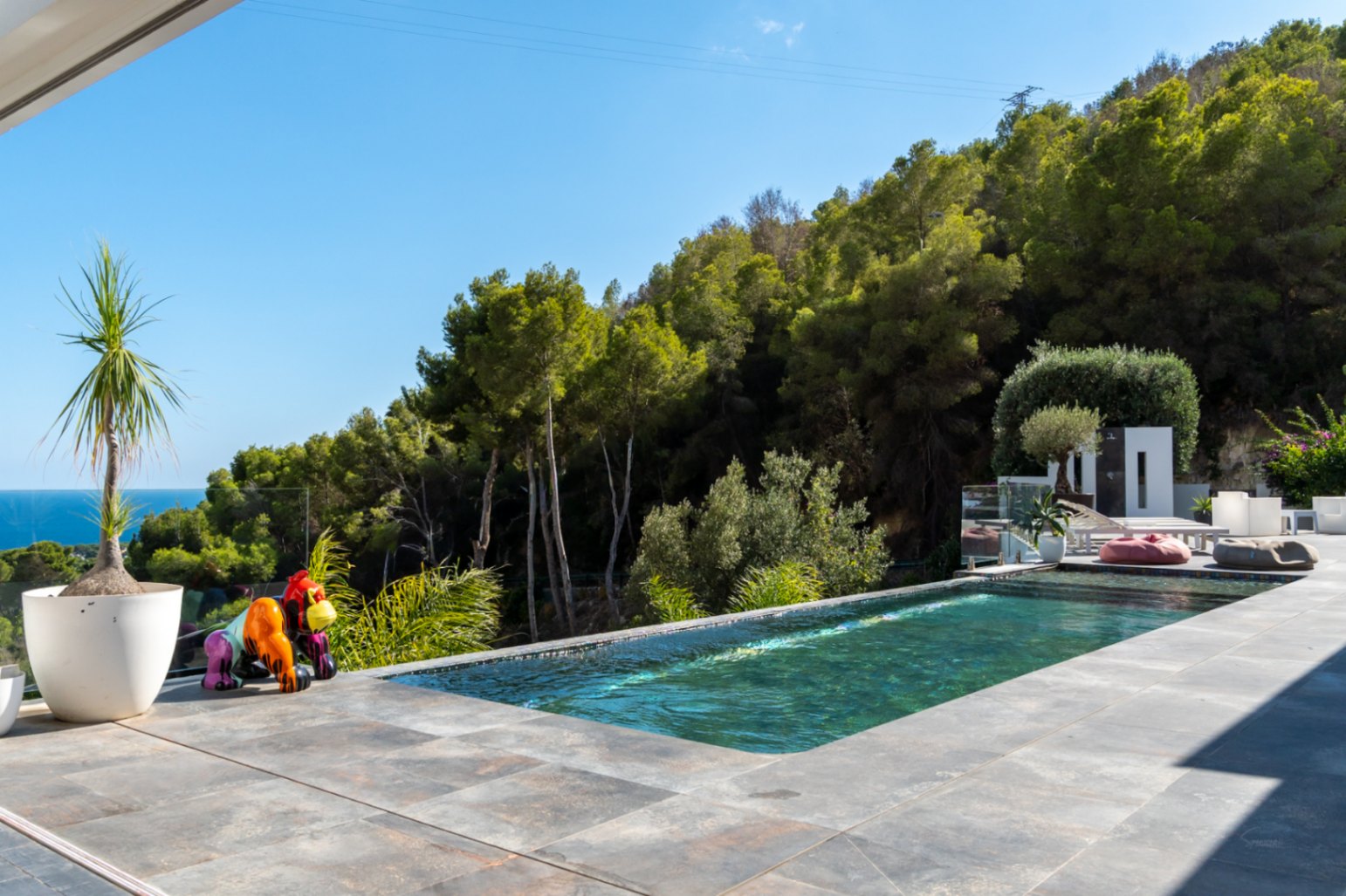 Infinity pool with turquoise water offering sea and mountain views, featuring a large-format stone terrace and a vibrant pop-art sculpture.