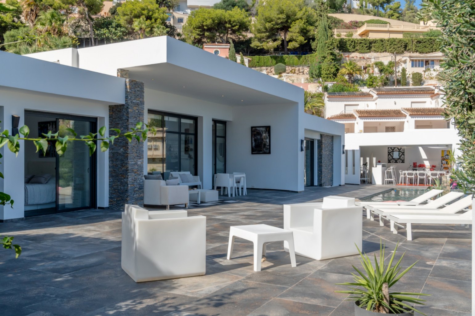 Main terrace featuring dark stone tiling, minimalist white lounge furniture, and stone-clad accents on the white facade near the pool area.