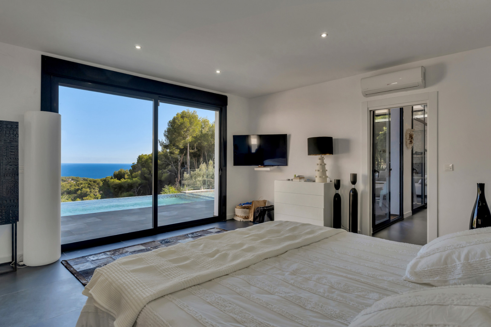 Master bedroom featuring floor-to-ceiling sliding glass doors with views of the infinity pool and Mediterranean Sea. Equipped with recessed lighting and dark tiled floors.