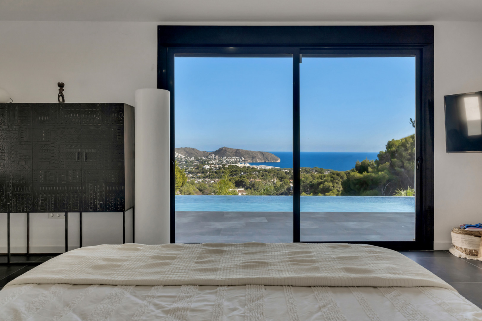 Master bedroom featuring panoramic sea and coastal views through large sliding glass doors. The space offers direct access to the infinity pool and terrace.